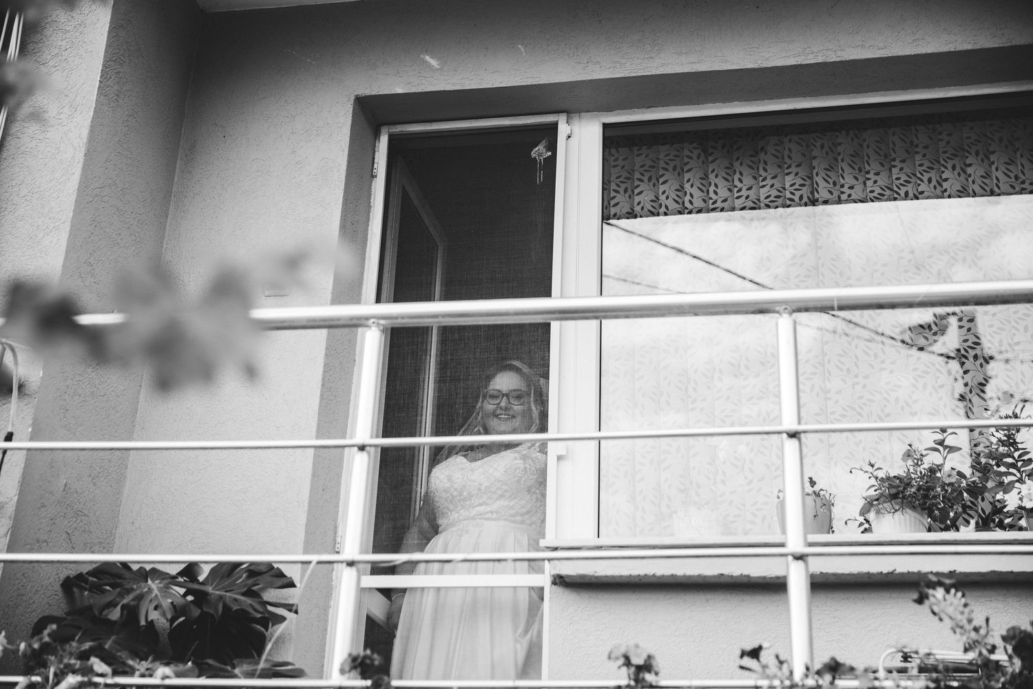 Black and white photo of a bride in a white dress standing at an open window on a building balcony.