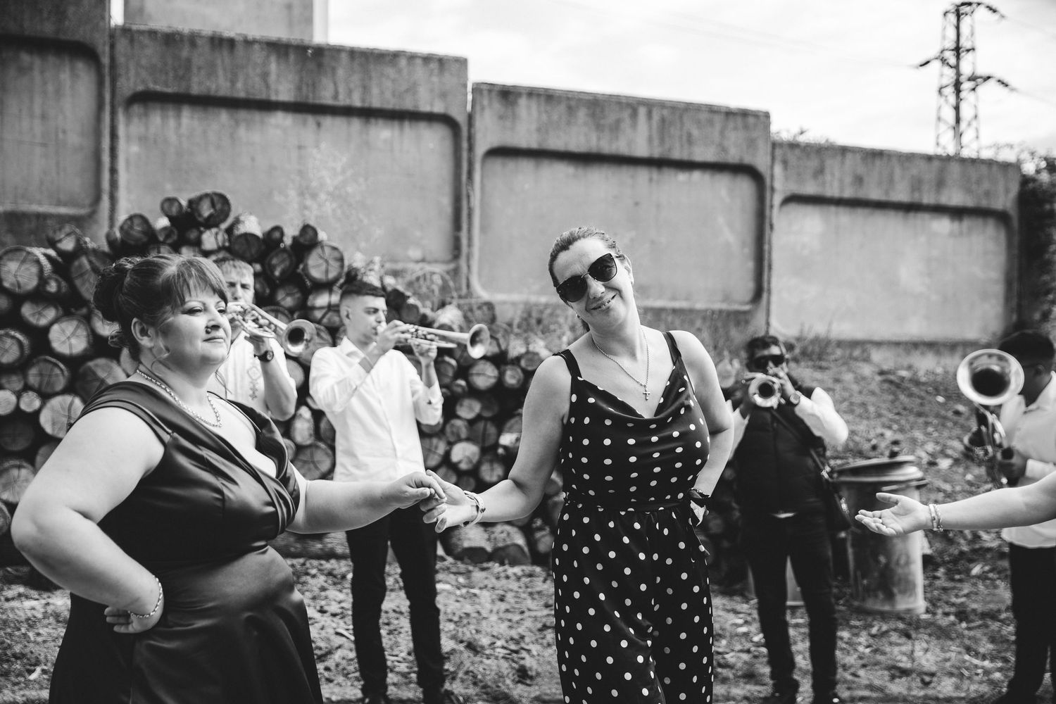 Black and white photo of people dancing outdoors near a live brass band with logs and a wall in the background.