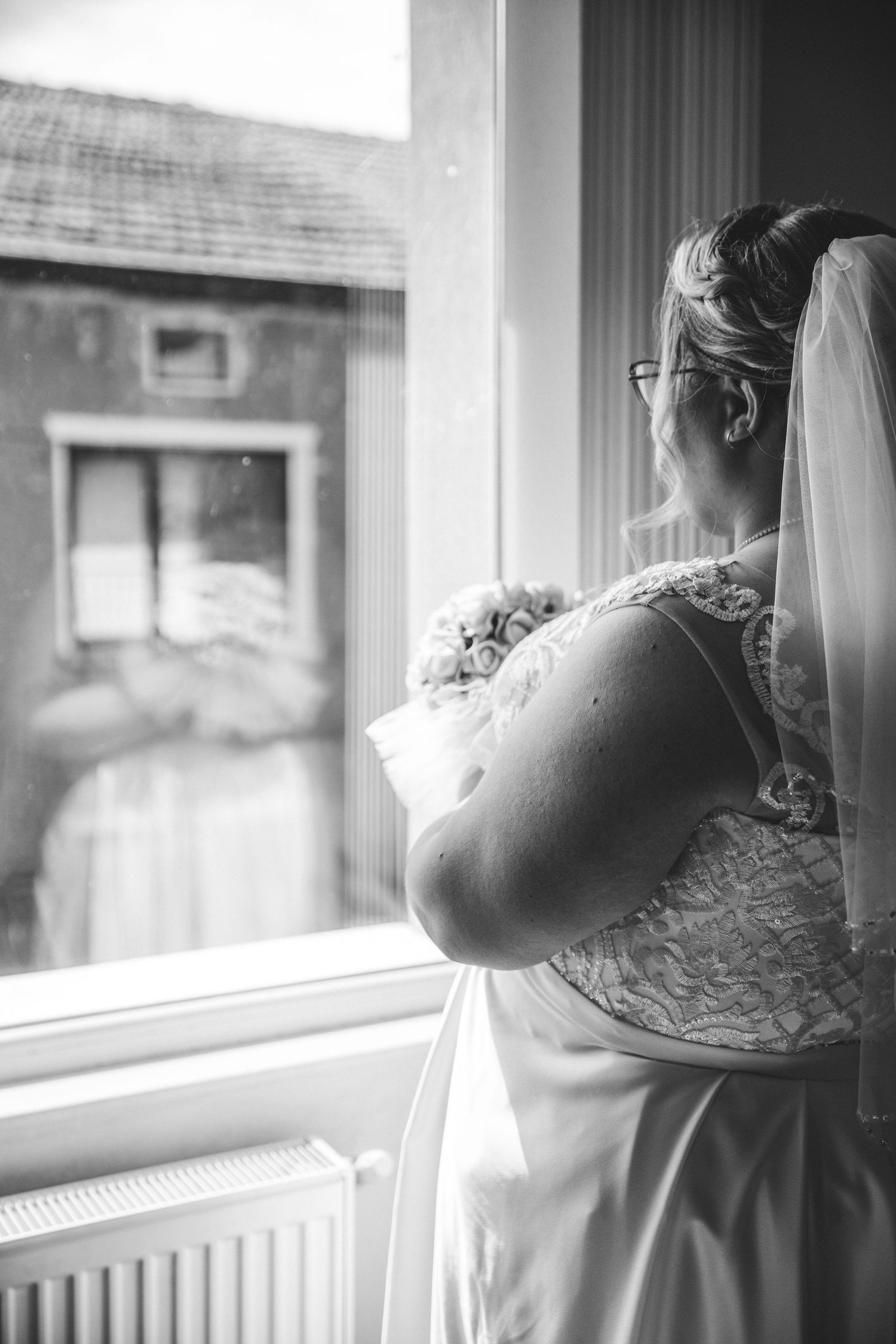 Black and white photo of a bride in lace gown and veil holding bouquet, gazing out a window on her wedding day.