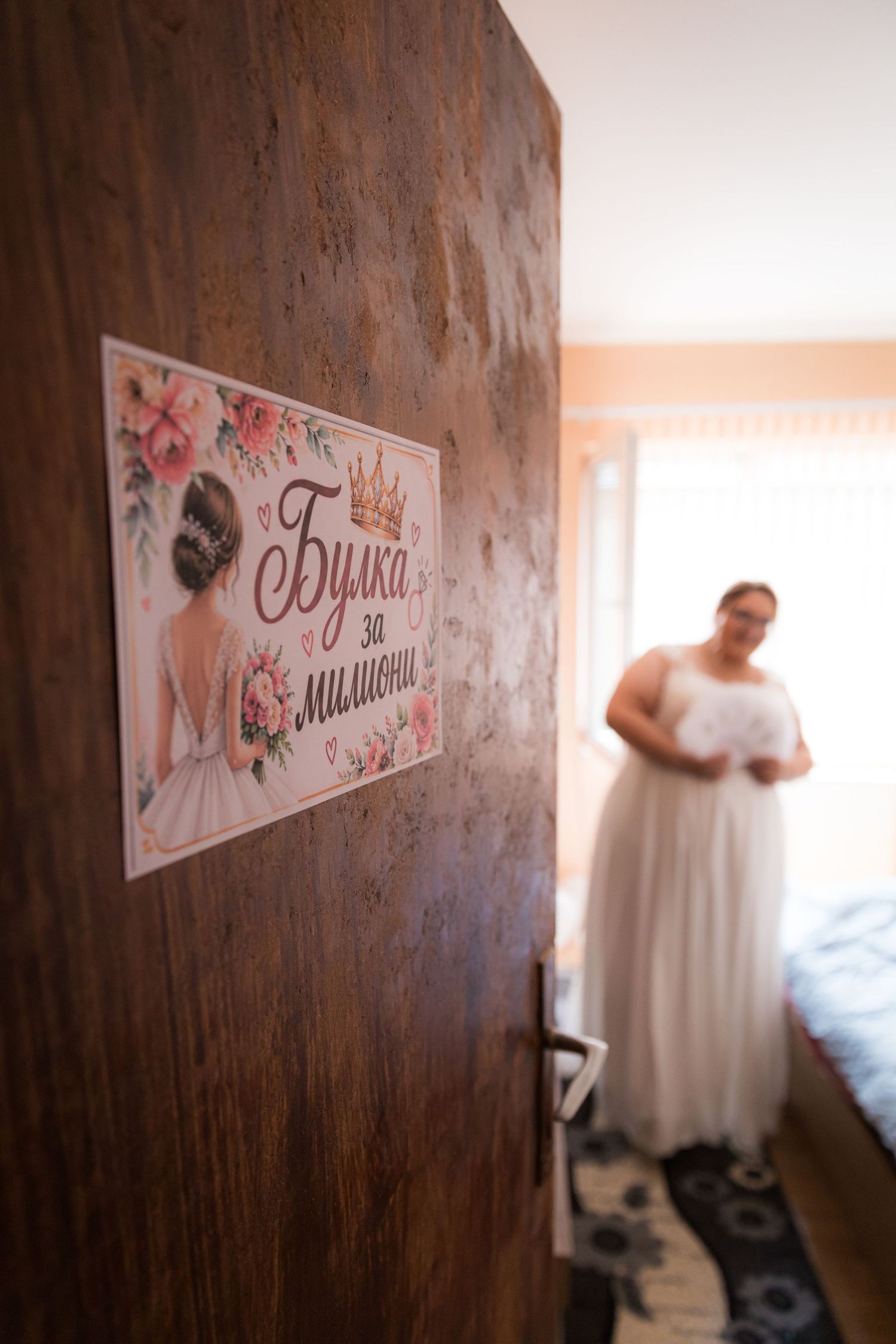A bride in a white wedding gown stands in a room behind a wooden door with a decorative bridal sign in Cyrillic.