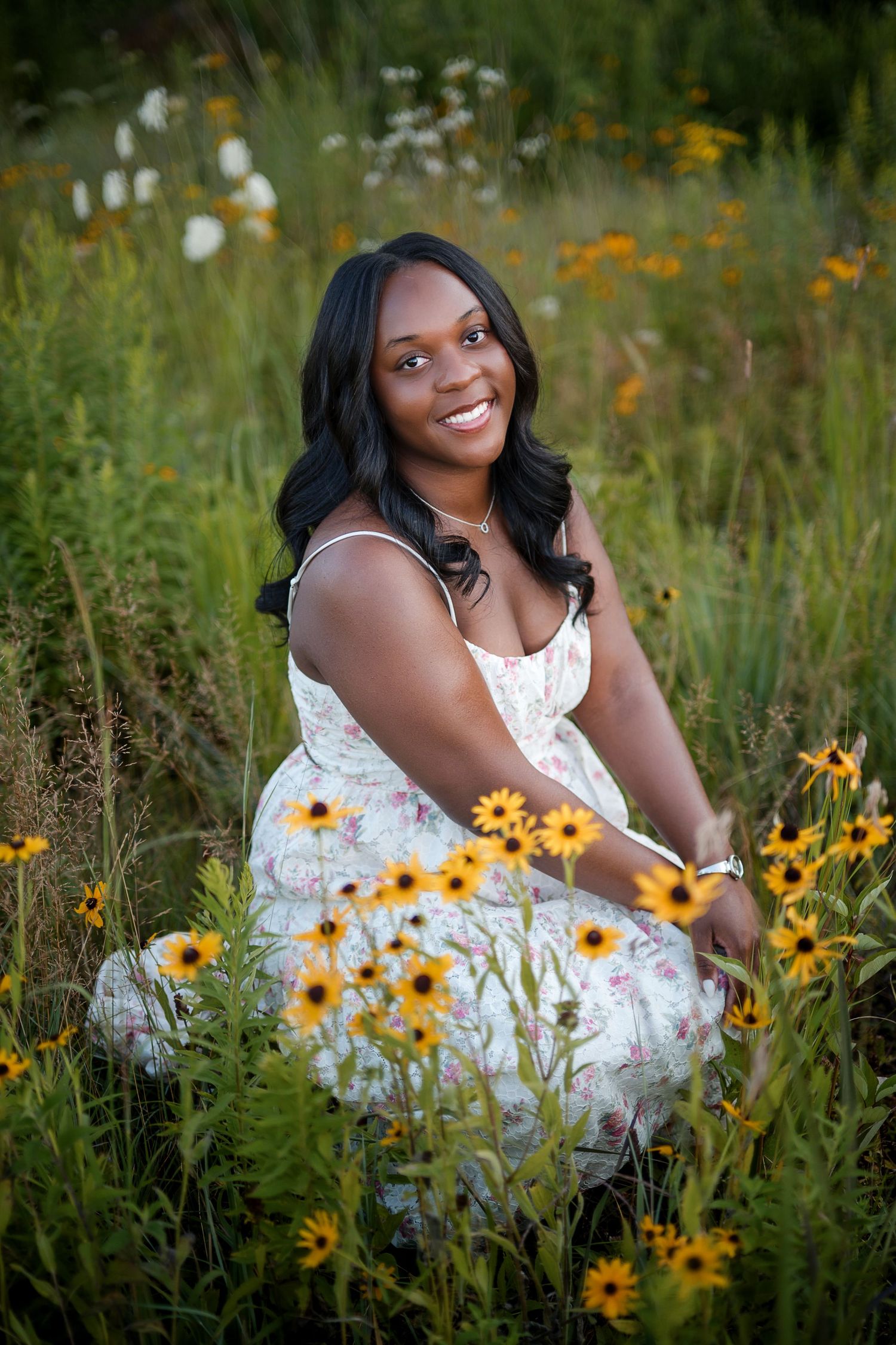 Young woman in floral dress sitting among wildflowers and black-eyed Susans, smiling outdoors.