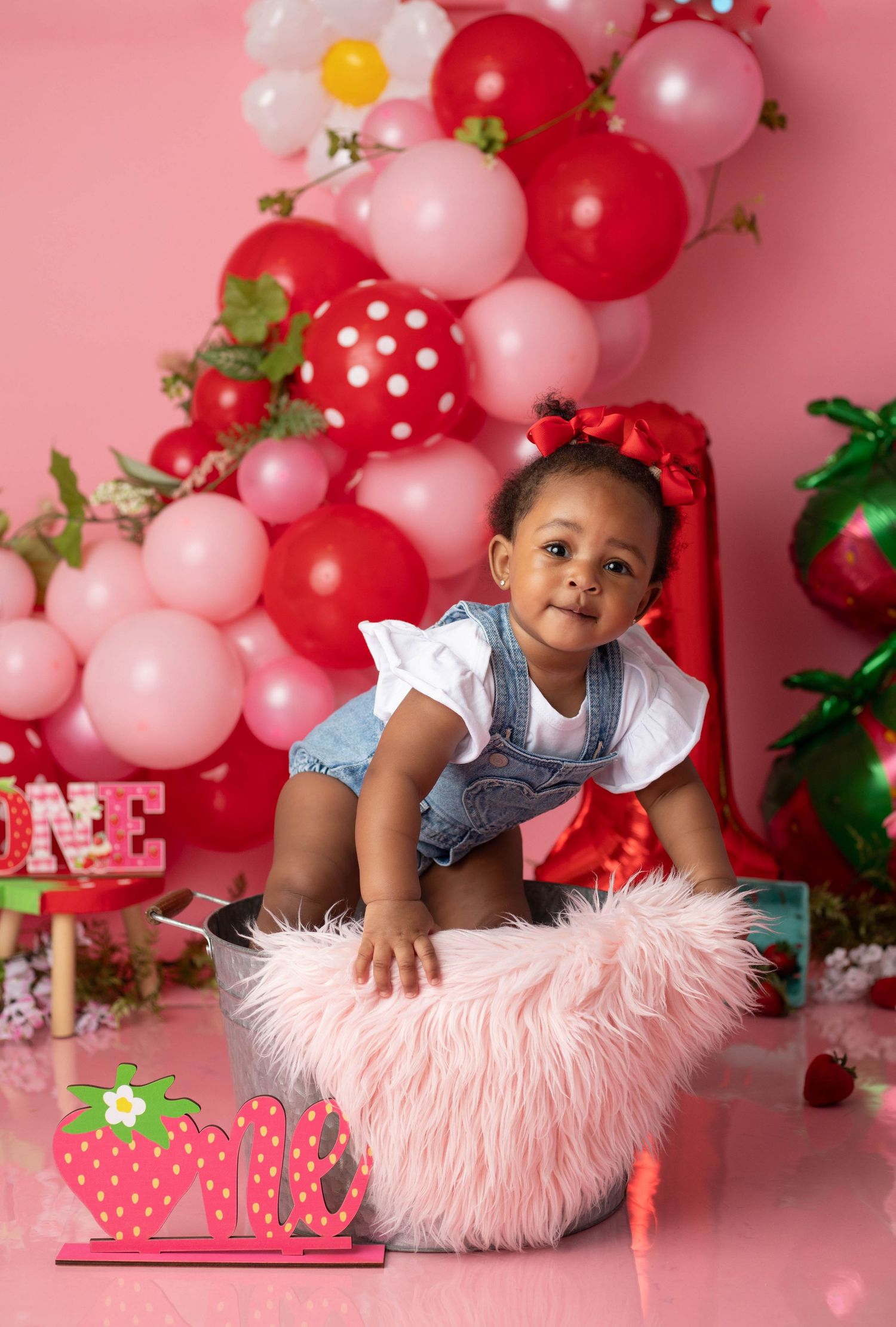 Baby girl in denim overalls sits in pink fluffy bowl at strawberry-themed first birthday cake smash with balloon backdrop.