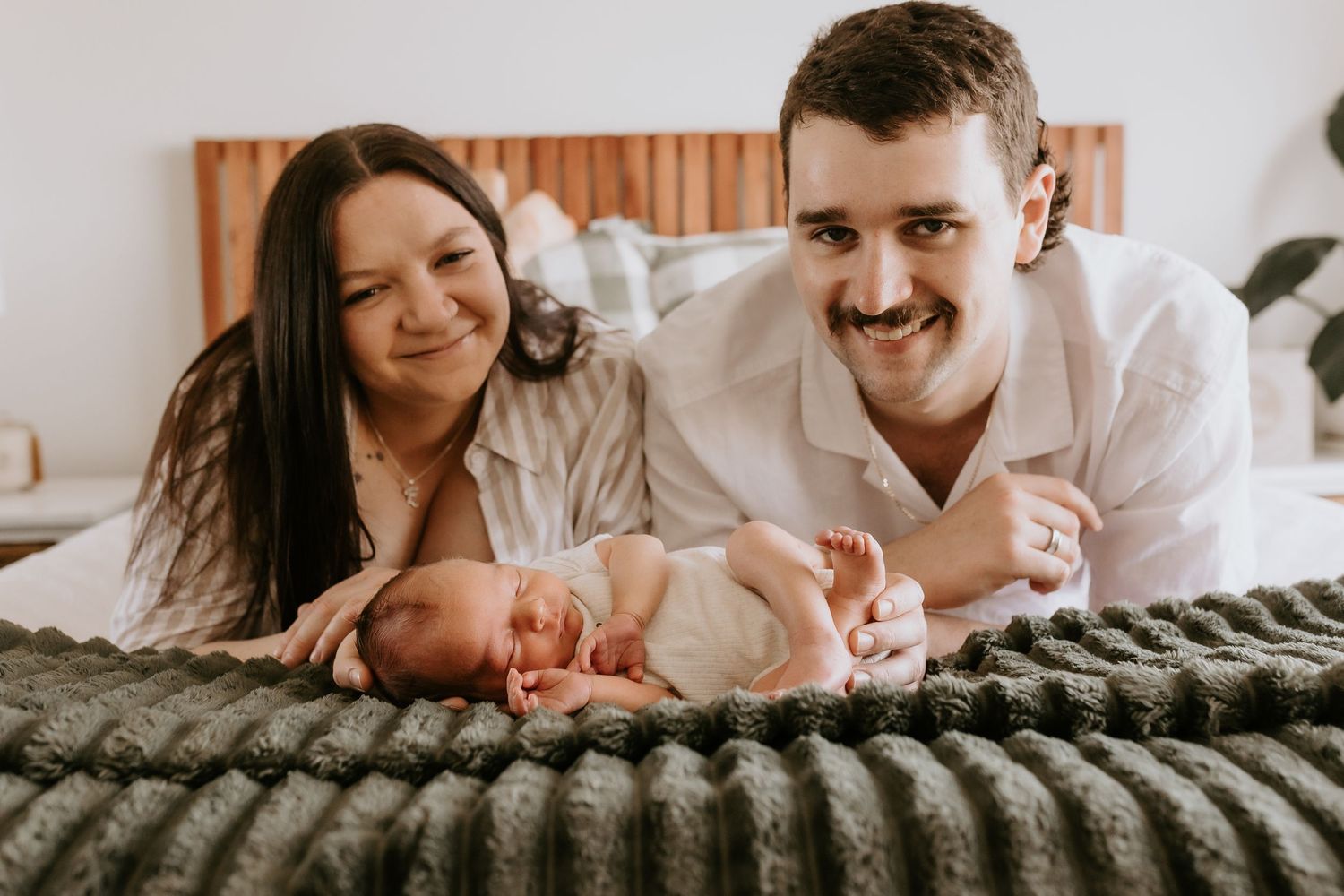 A happy couple smiles while lying on a bed with their newborn baby on a dark green blanket.