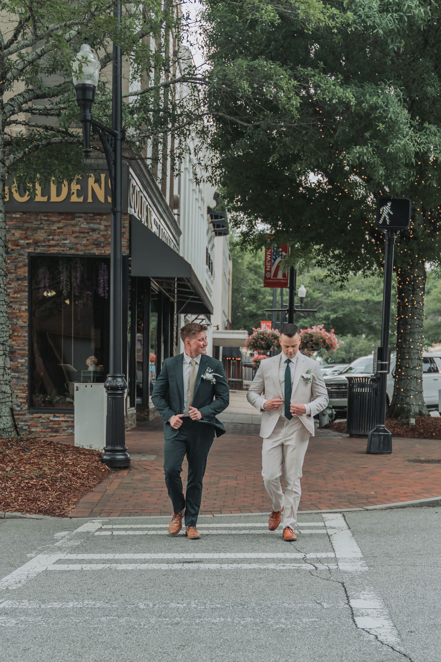 Two well-dressed groomsmen in suits walk across a crosswalk in a charming downtown shopping district.