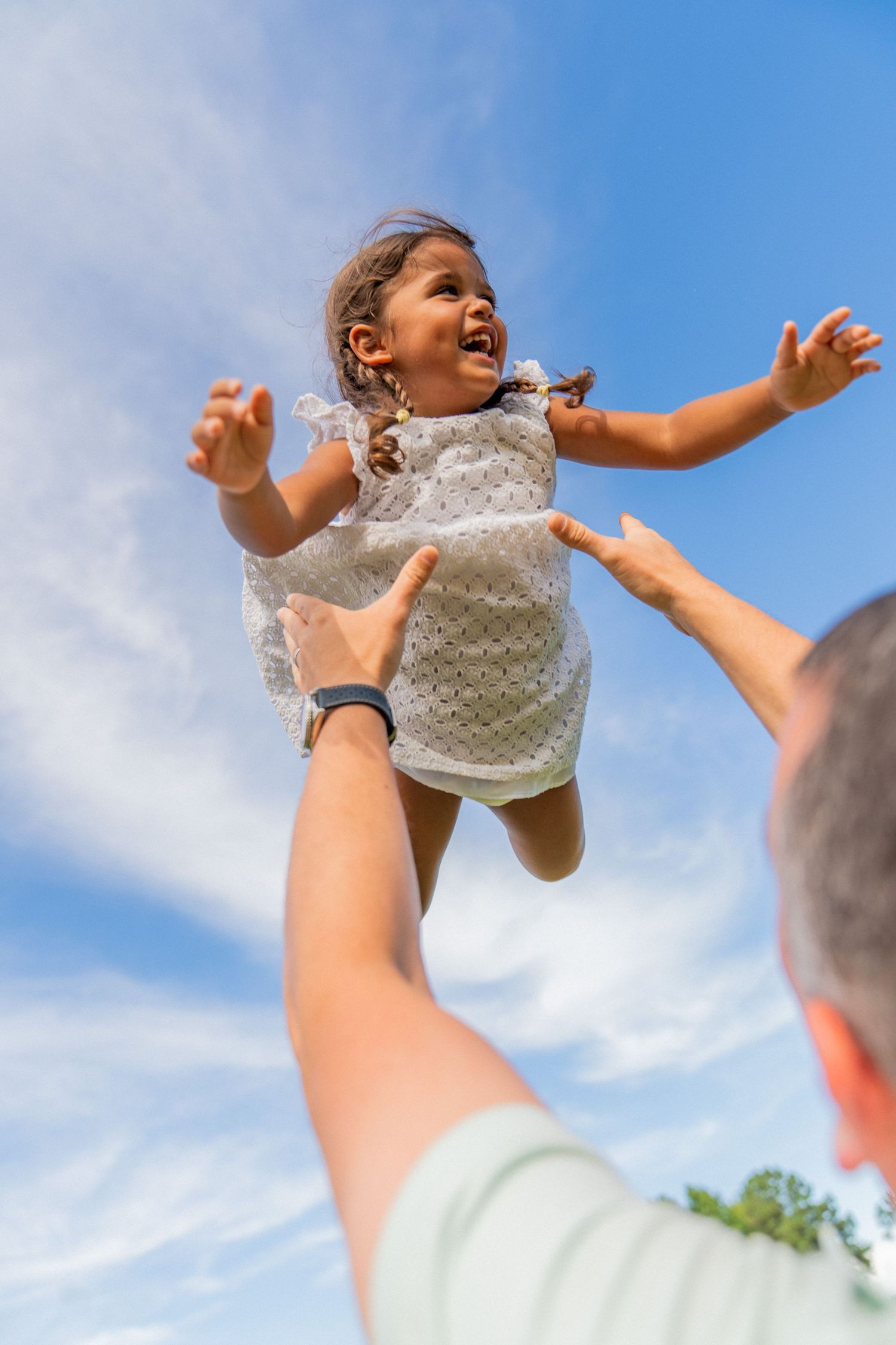 A joyful little girl in a white dress is tossed into the blue sky by her father's outstretched hands.