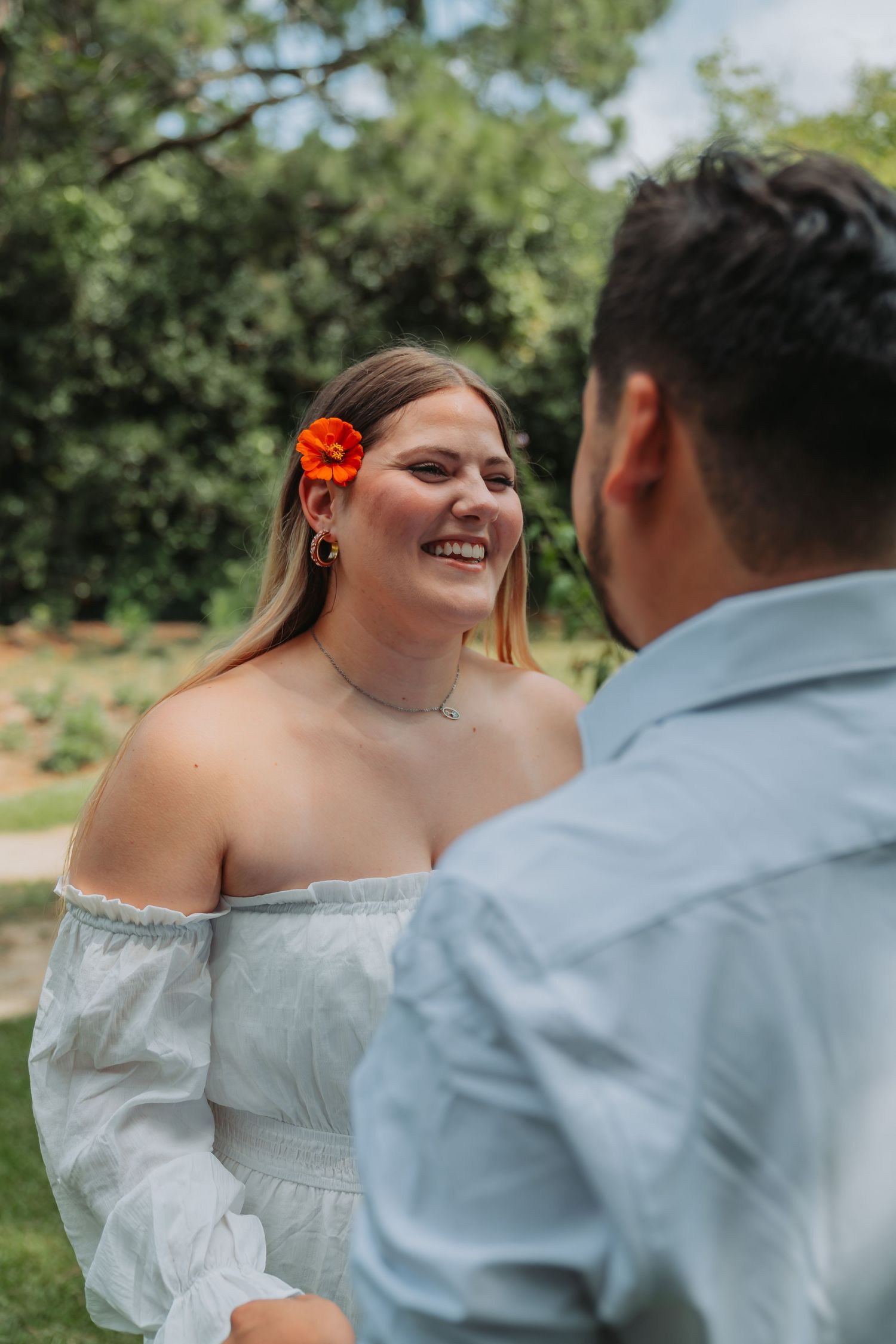 Smiling bride in white off-shoulder dress with orange flower in hair, holding hands with groom outdoors.