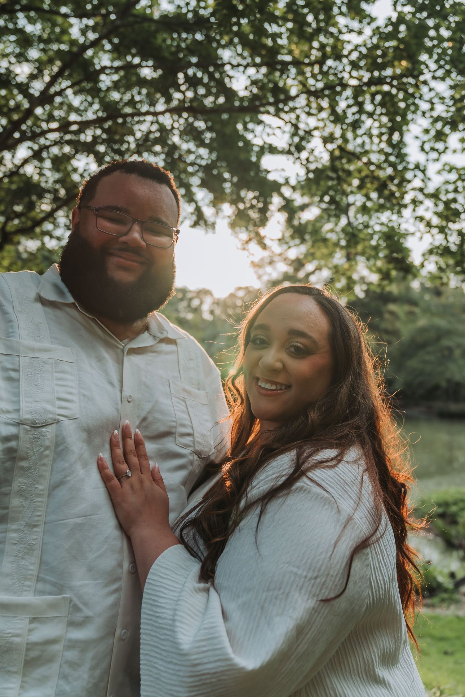 Couple smiling outdoors under green trees at golden hour, woman resting hand on mans chest, both wearing white.