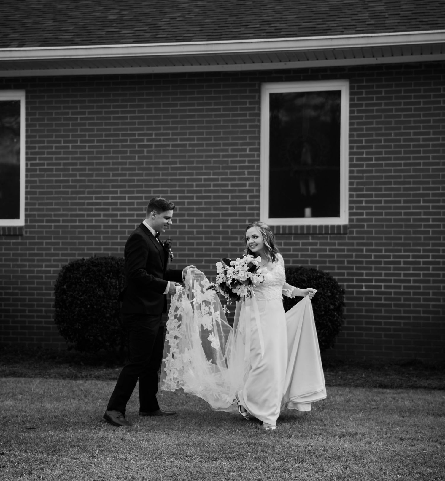 Black and white photo of a bride and groom walking outside a brick church, groom holding the brides lace veil train.