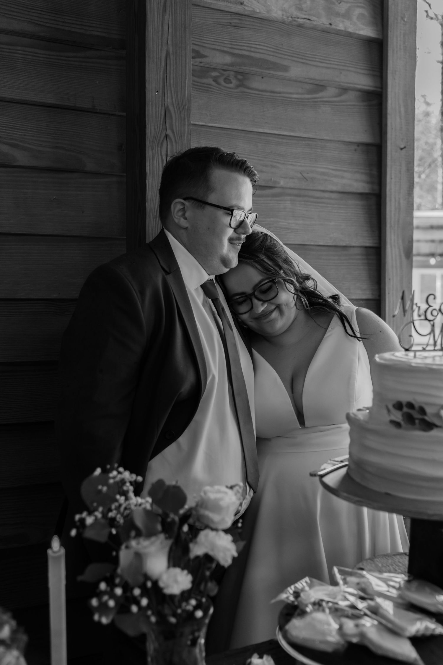 Black and white photo of a smiling bride and groom leaning together near their wedding cake at a rustic barn reception.
