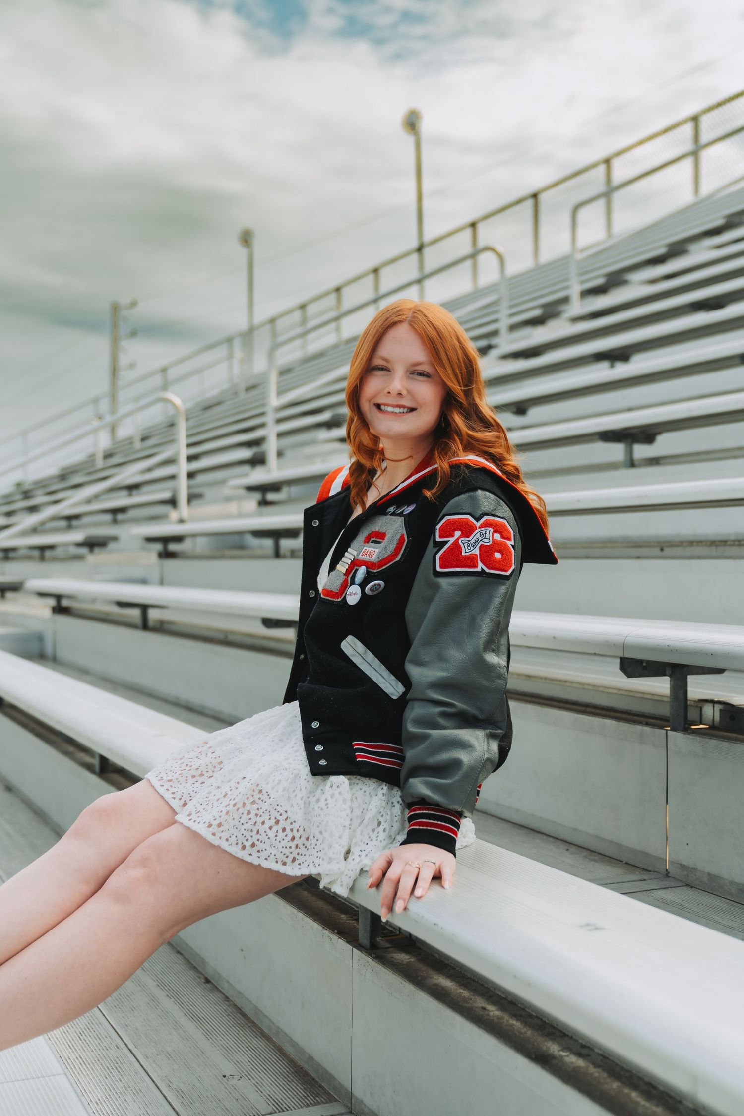 Smiling redhead teen in varsity jacket #23 poses on bleachers at a stadium, wearing a white lace skirt.