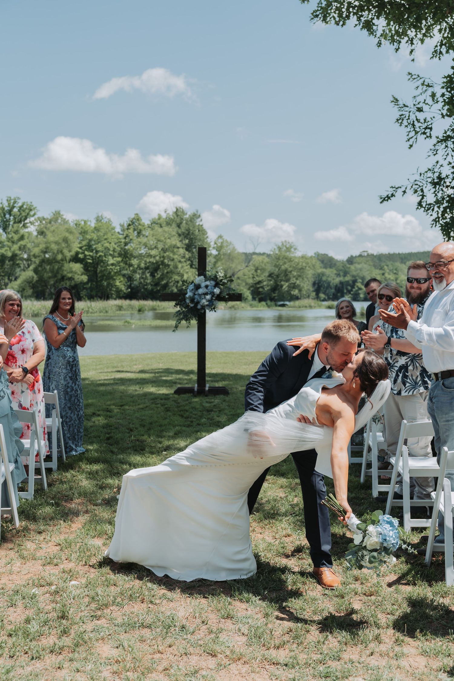 Groom dips bride for a kiss at outdoor wedding ceremony by a lake, guests applauding in background on sunny day.