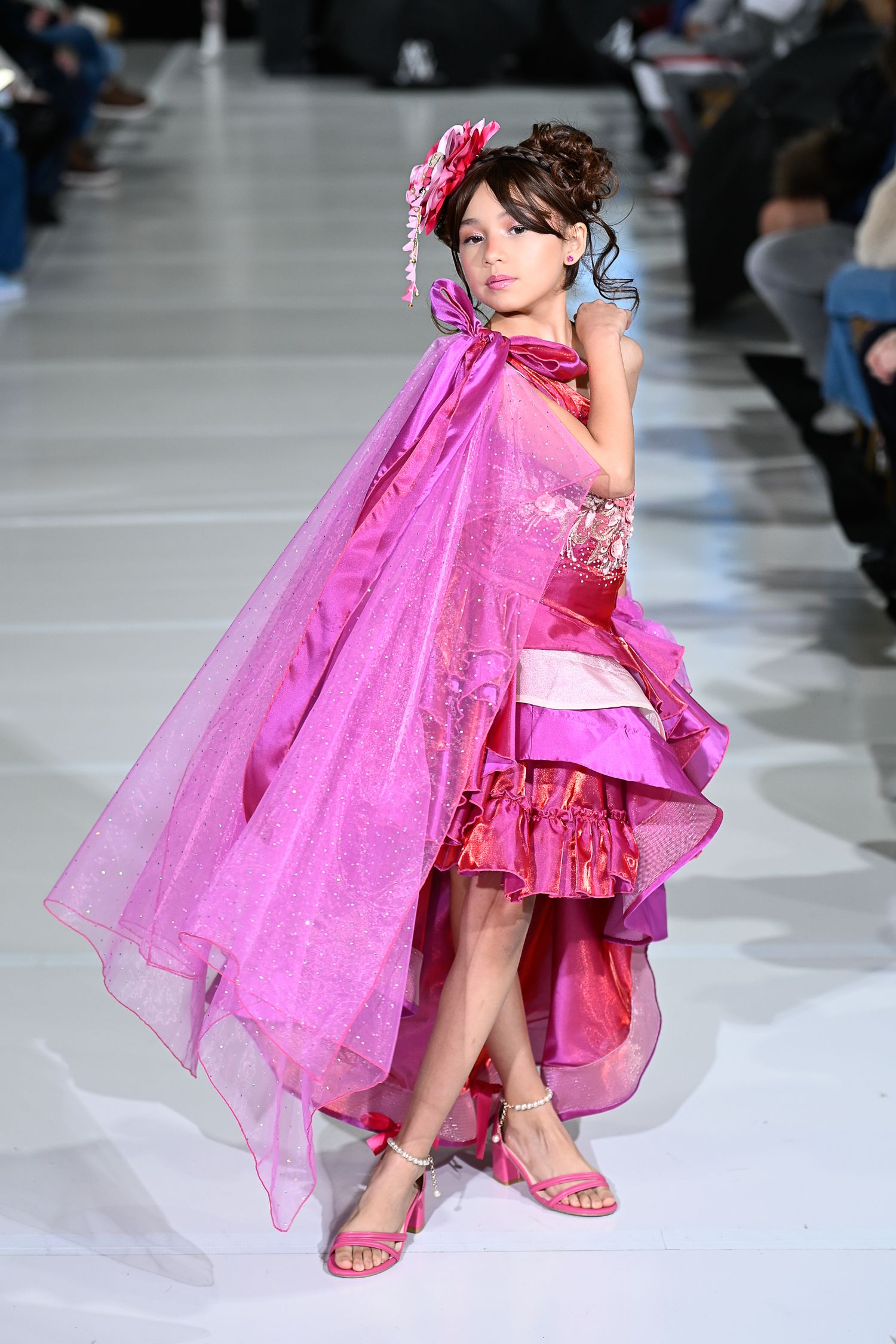 Young girl models a vibrant pink layered gown with flowing cape on a fashion runway.