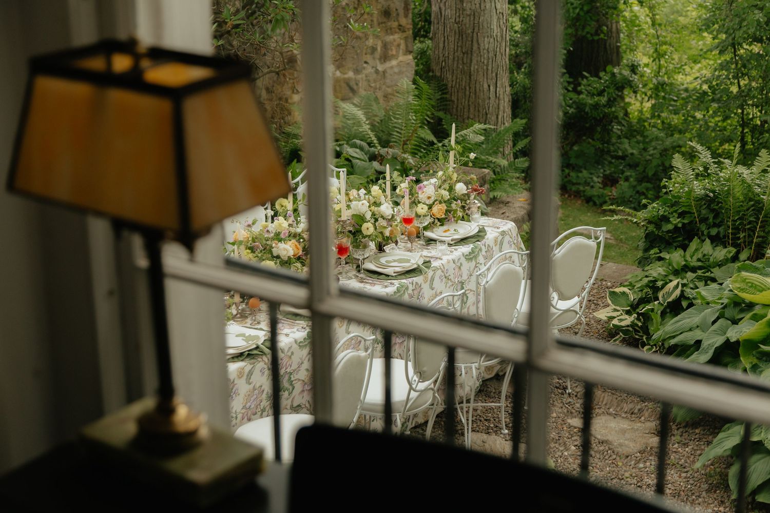 Elegant outdoor garden dining table with floral centerpiece viewed through a window, with a vintage lamp in the foreground.