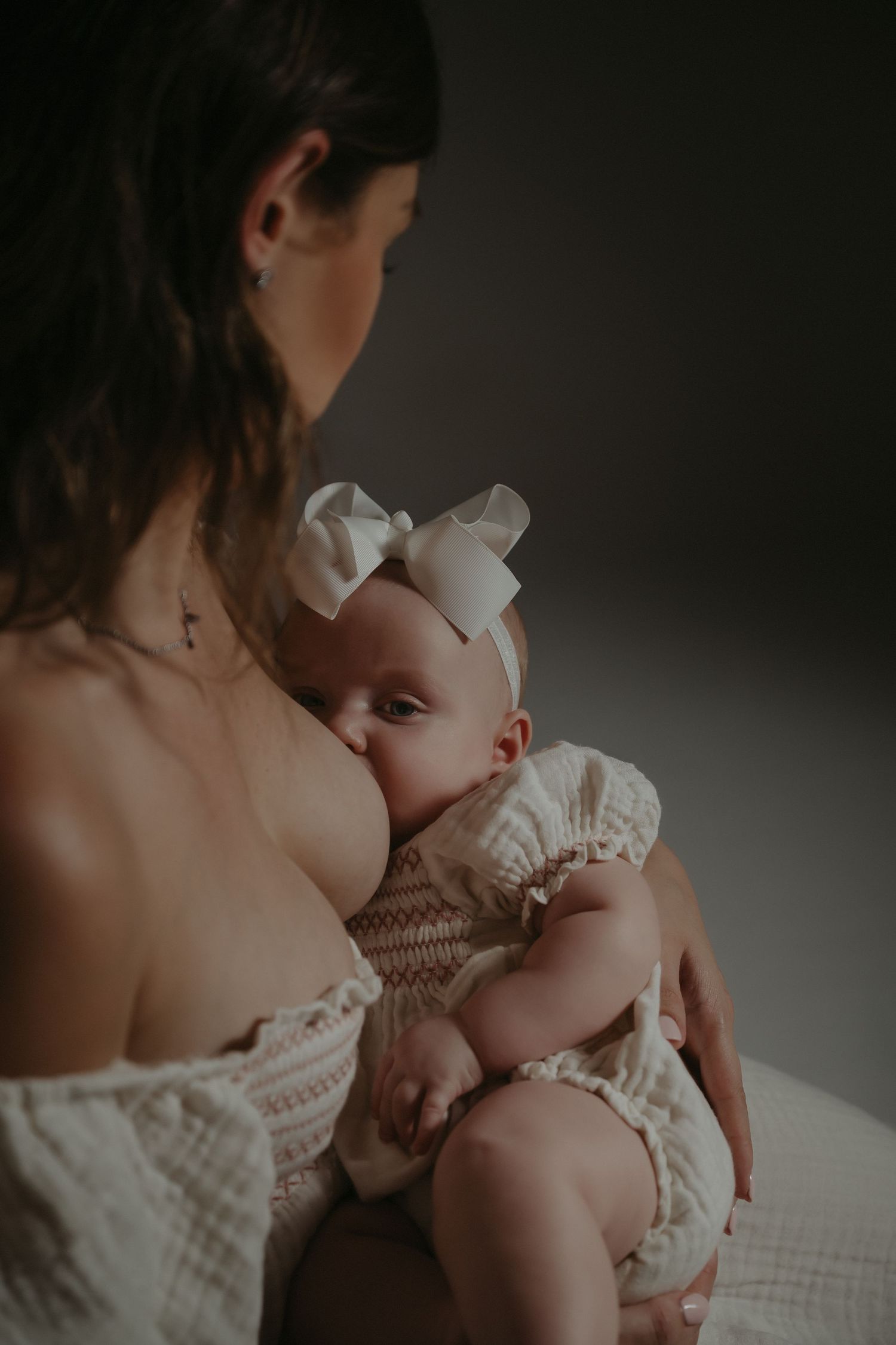 A mother breastfeeding her newborn baby girl wearing a white bow headband and smocked romper in a soft studio setting.