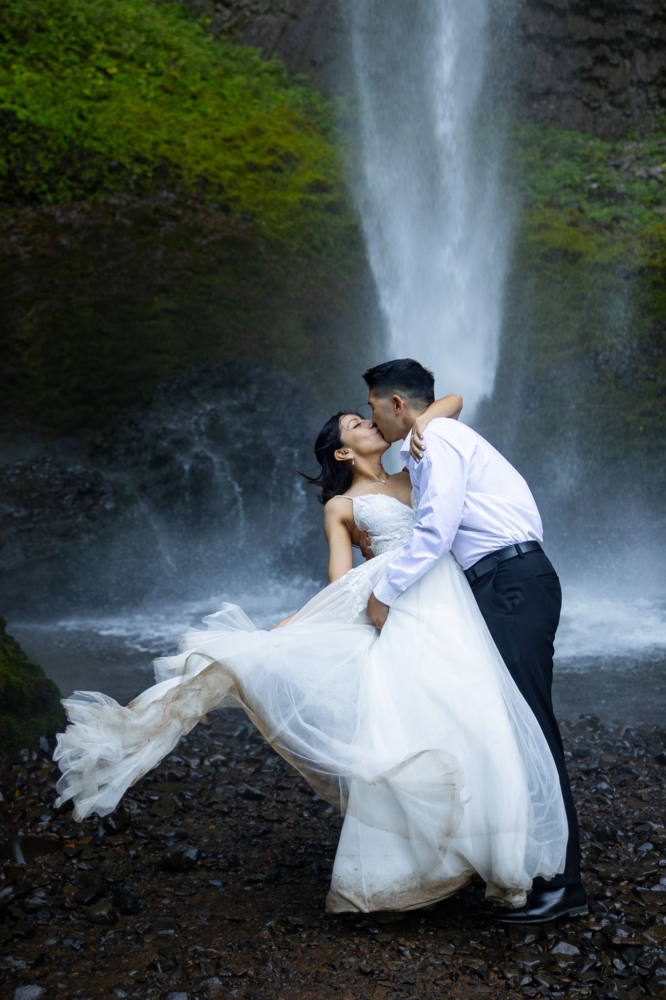 Bride and groom share a romantic kiss in front of a stunning waterfall, her white gown flowing in the breeze.