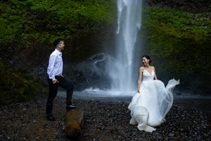 Couple poses before a waterfall, bride in flowing white gown, groom in white shirt, surrounded by lush greenery.