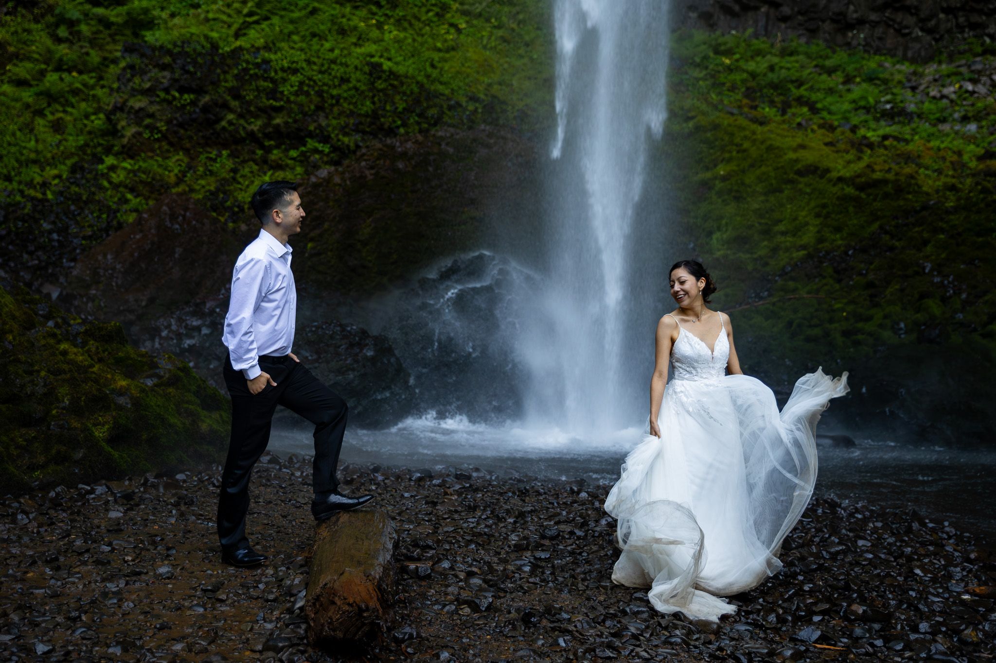 Couple poses before a waterfall, bride in flowing white gown, groom in white shirt, surrounded by lush greenery.