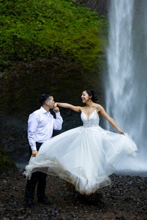 Groom kisses bride's hand as she twirls her white gown near a cascading waterfall with green foliage backdrop.