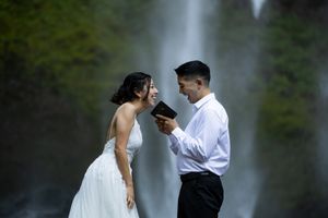 Couple in wedding attire laughing together in front of a waterfall, groom holding a book.