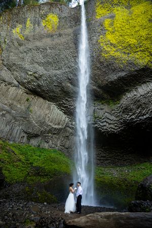 Bride and groom hold hands before a towering waterfall surrounded by lush green foliage and basalt rock columns.