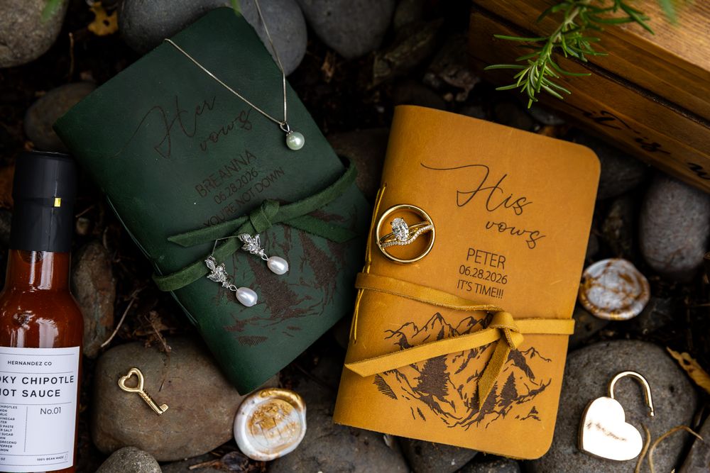 His and hers leather wedding journals with mountain engraving, rings, pearl earrings, and heart charm on rustic stone background.