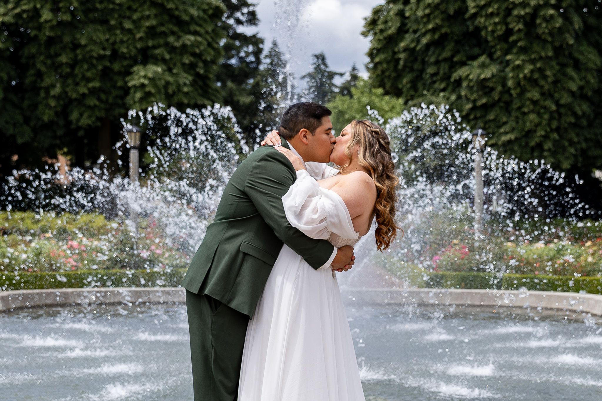 Bride and groom share a romantic kiss in front of a garden fountain on their wedding day.