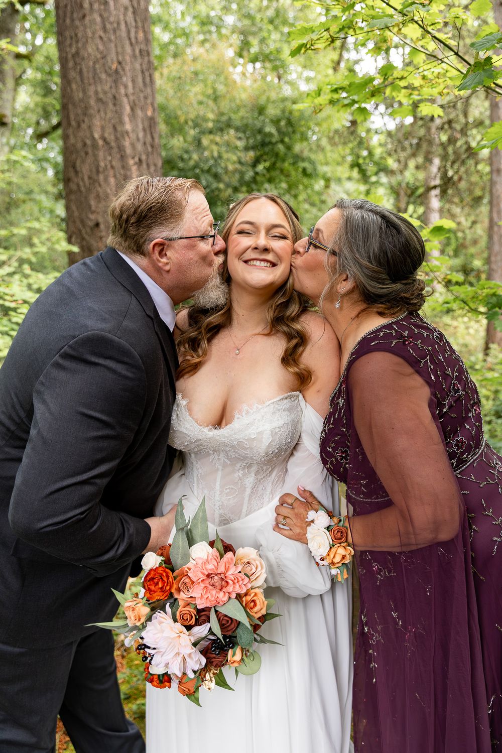 Smiling bride in white gown receives kisses from parents while holding a colorful bouquet outdoors.
