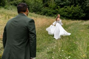 Bride in white gown twirling in meadow while groom in green suit watches during outdoor wedding photo session.