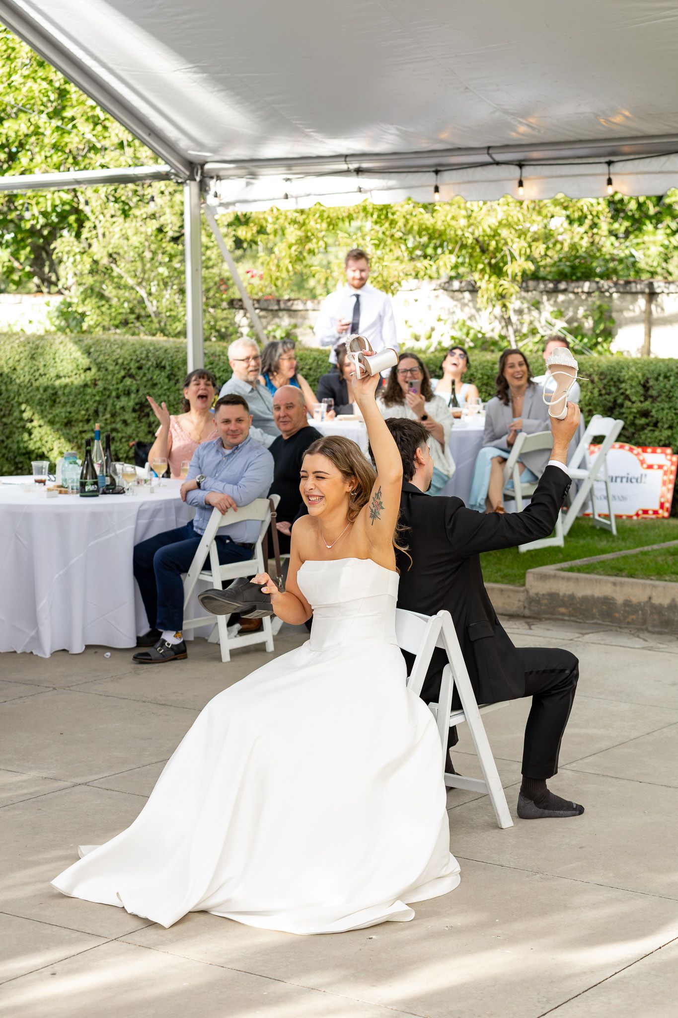 Bride and groom celebrate joyfully at outdoor wedding reception with smiling guests seated under white tent.