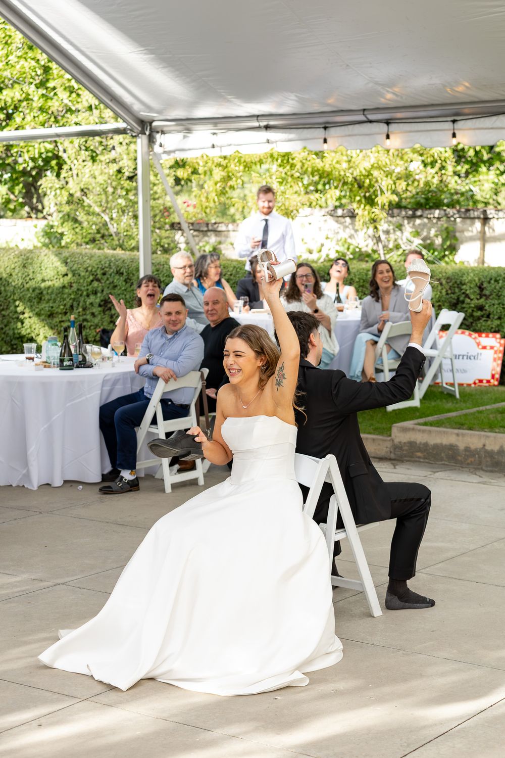 Bride and groom celebrate joyfully at outdoor wedding reception with smiling guests seated under white tent.