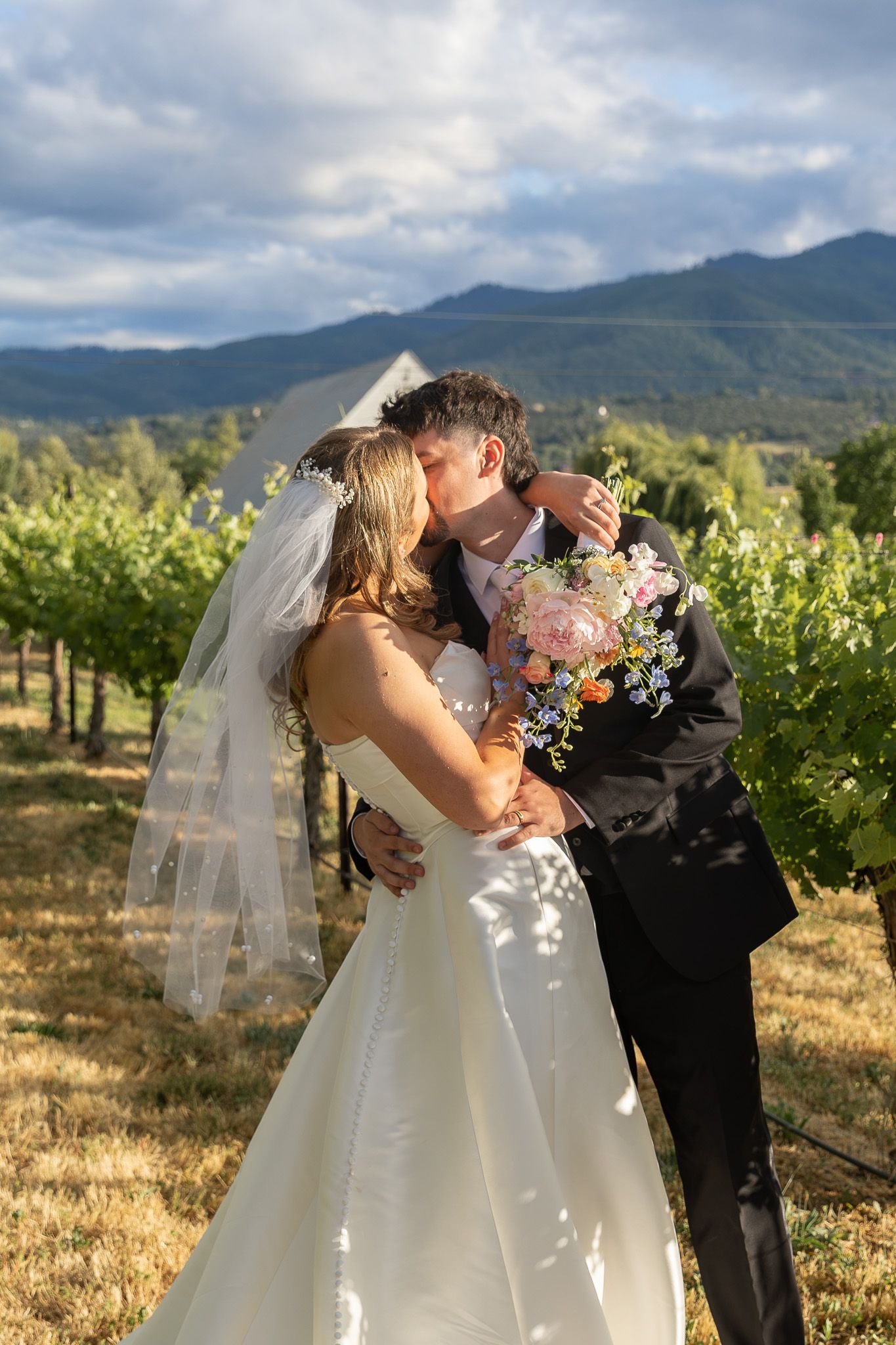 Bride and groom kissing in a vineyard, bride in white gown with veil holding floral bouquet, mountains in background.