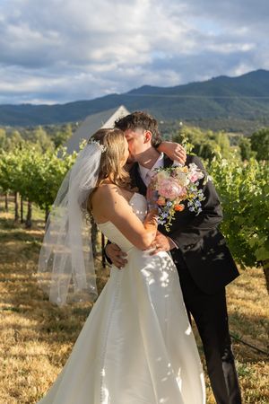 Bride and groom kissing in a vineyard, bride in white gown with veil holding floral bouquet, mountains in background.