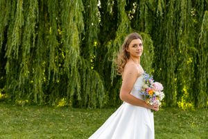 Bride in white gown holding colorful bouquet stands before weeping willow tree on sunny day.