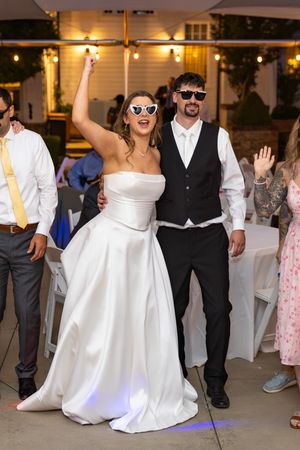 Bride in white gown and groom in vest celebrate at outdoor wedding reception wearing heart-shaped sunglasses.