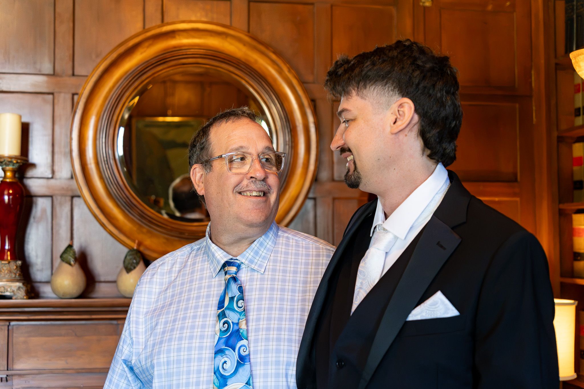 Two men in formal attire smiling at each other in an elegantly decorated room with wood paneling and a round mirror.