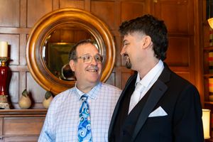 Two men in formal attire smiling at each other in an elegantly decorated room with wood paneling and a round mirror.
