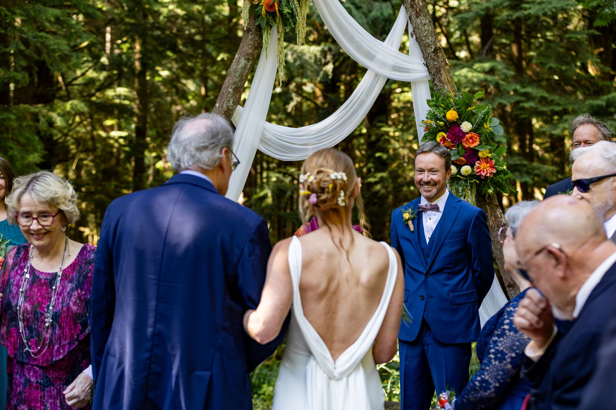 Outdoor forest wedding ceremony with bride in backless gown walking toward smiling groom under floral arch.