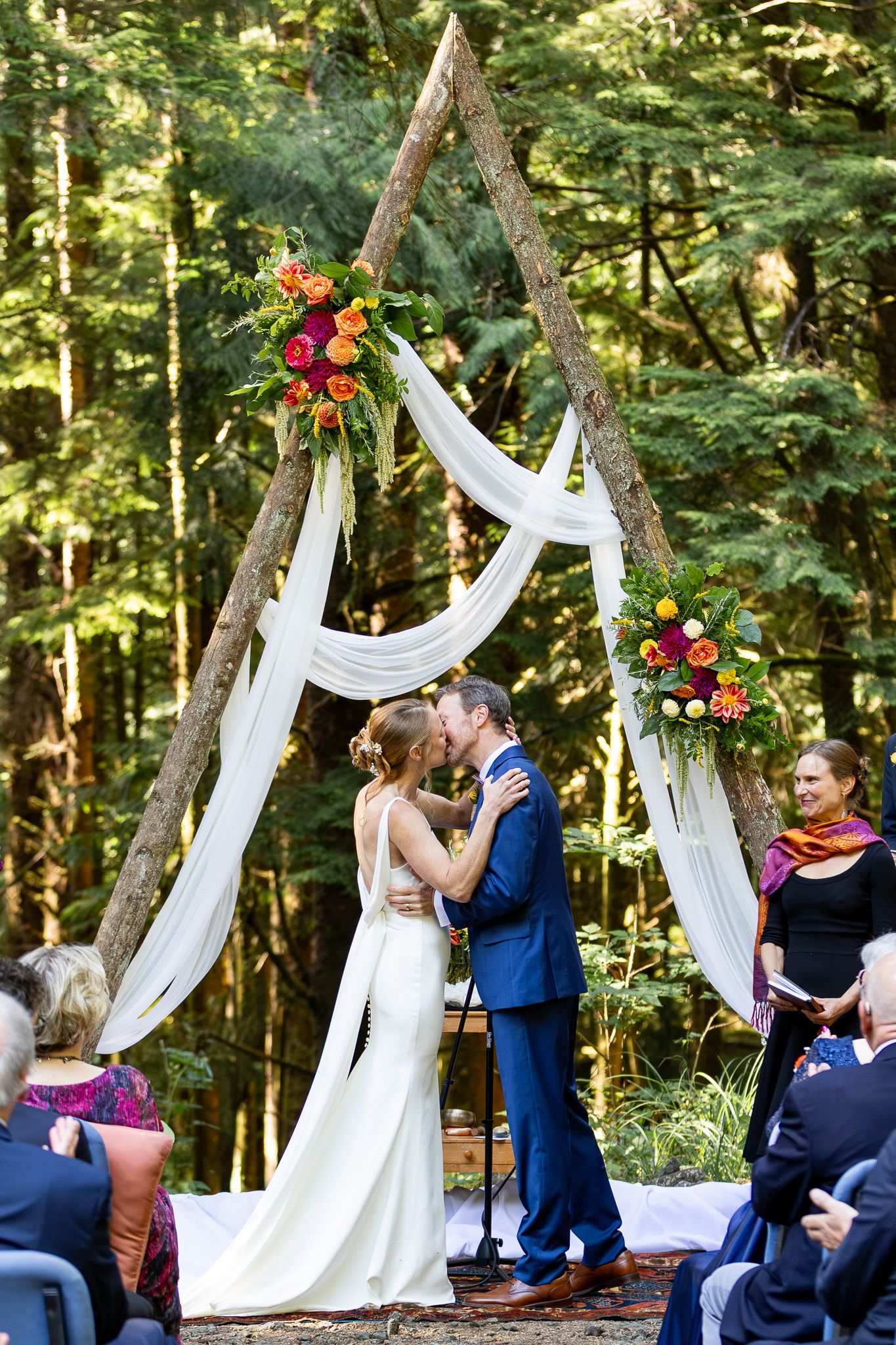 Bride and groom share first kiss under rustic triangular wooden arch with white draping and colorful florals in forest wedding.