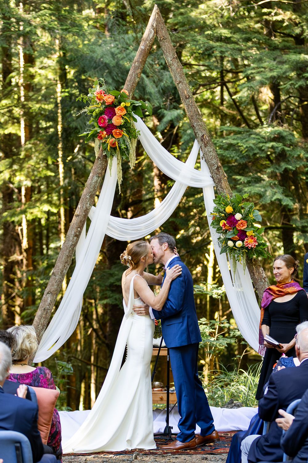 Bride and groom share first kiss under rustic triangular wooden arch with white draping and colorful florals in forest wedding.