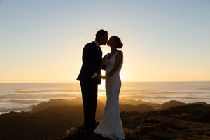 Silhouette of a couple kissing at sunset on rocky coastal cliffs, bride in white gown, groom in suit.