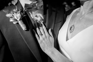 Black and white close-up of bride and groom hands showing wedding rings with boutonniere and necklace details.