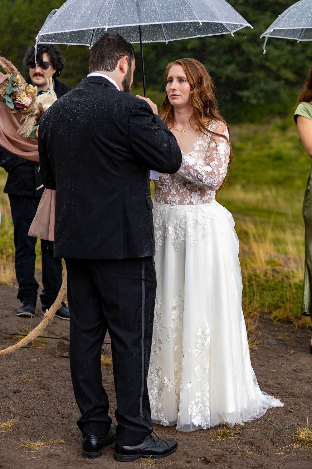 Bride in lace wedding dress exchanges vows with groom in black suit during outdoor rainy ceremony with guests holding umbrellas.