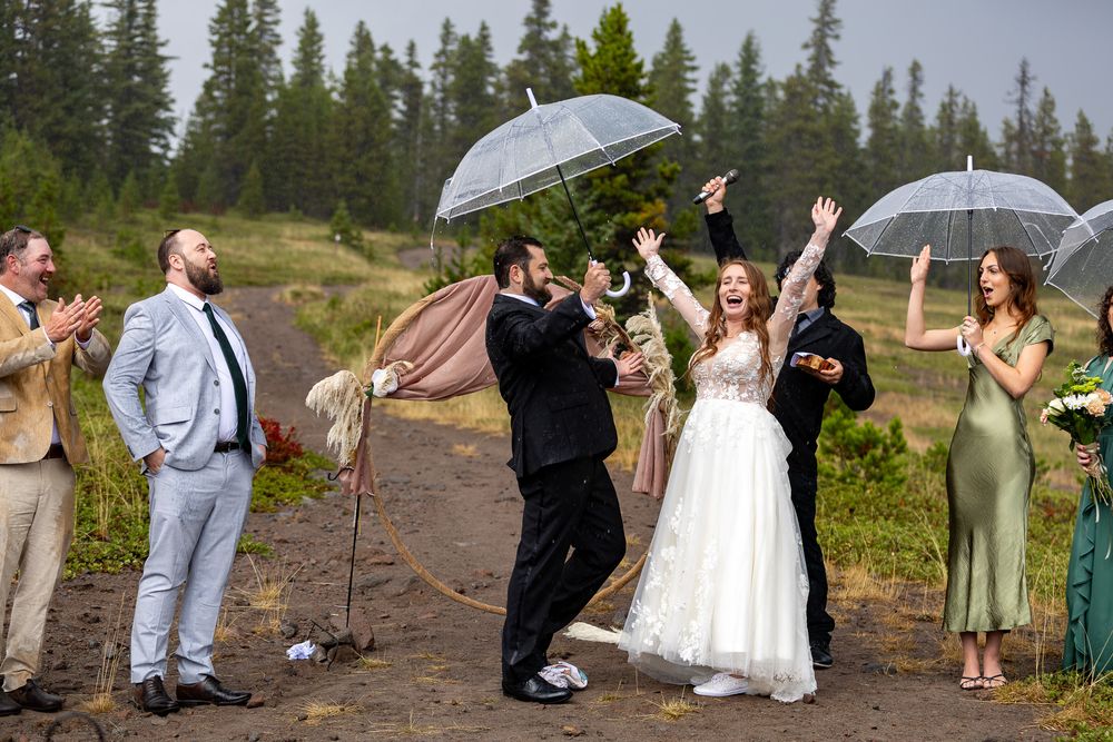 A joyful bride and groom celebrate outdoors in the rain with guests holding clear umbrellas at a rustic mountain wedding.