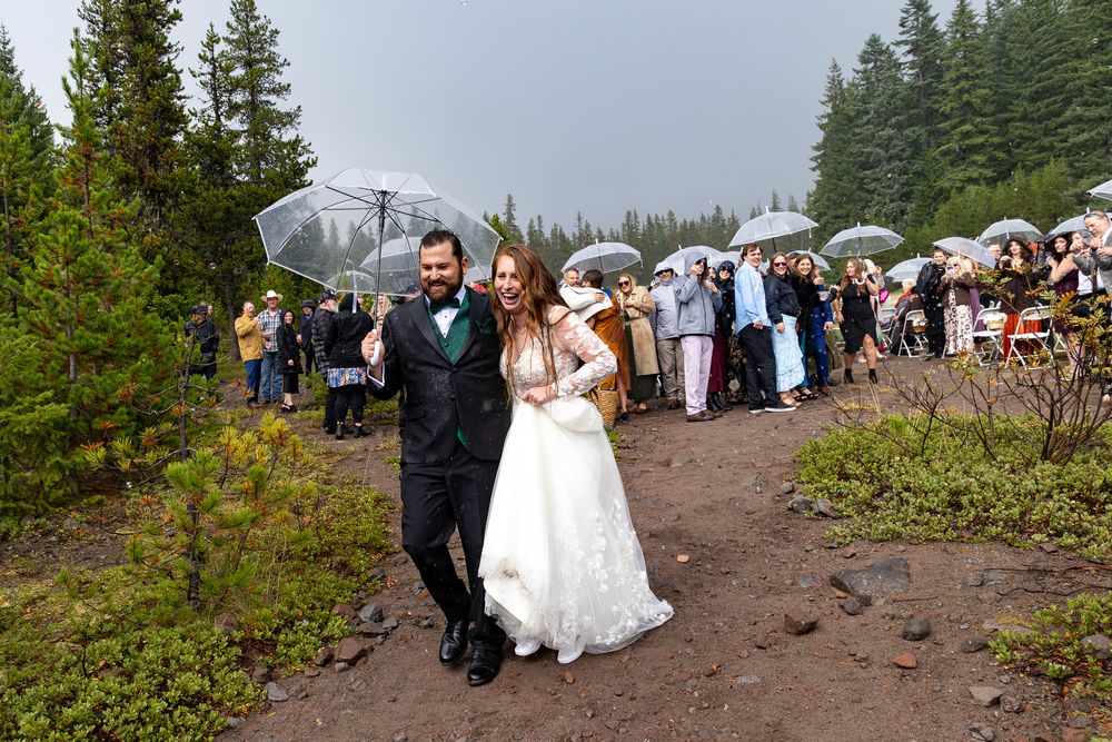 Bride and groom walk together outdoors at rainy forest wedding ceremony with guests holding umbrellas in background.