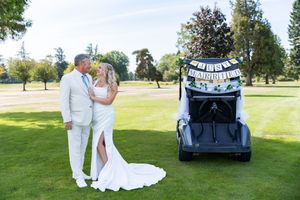 Newlywed couple in white attire posing beside a golf cart with Just Married banner on a lush green golf course.