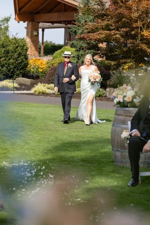 Bride in white gown walks down outdoor aisle with escort at sunny garden wedding ceremony near wine barrel decor.