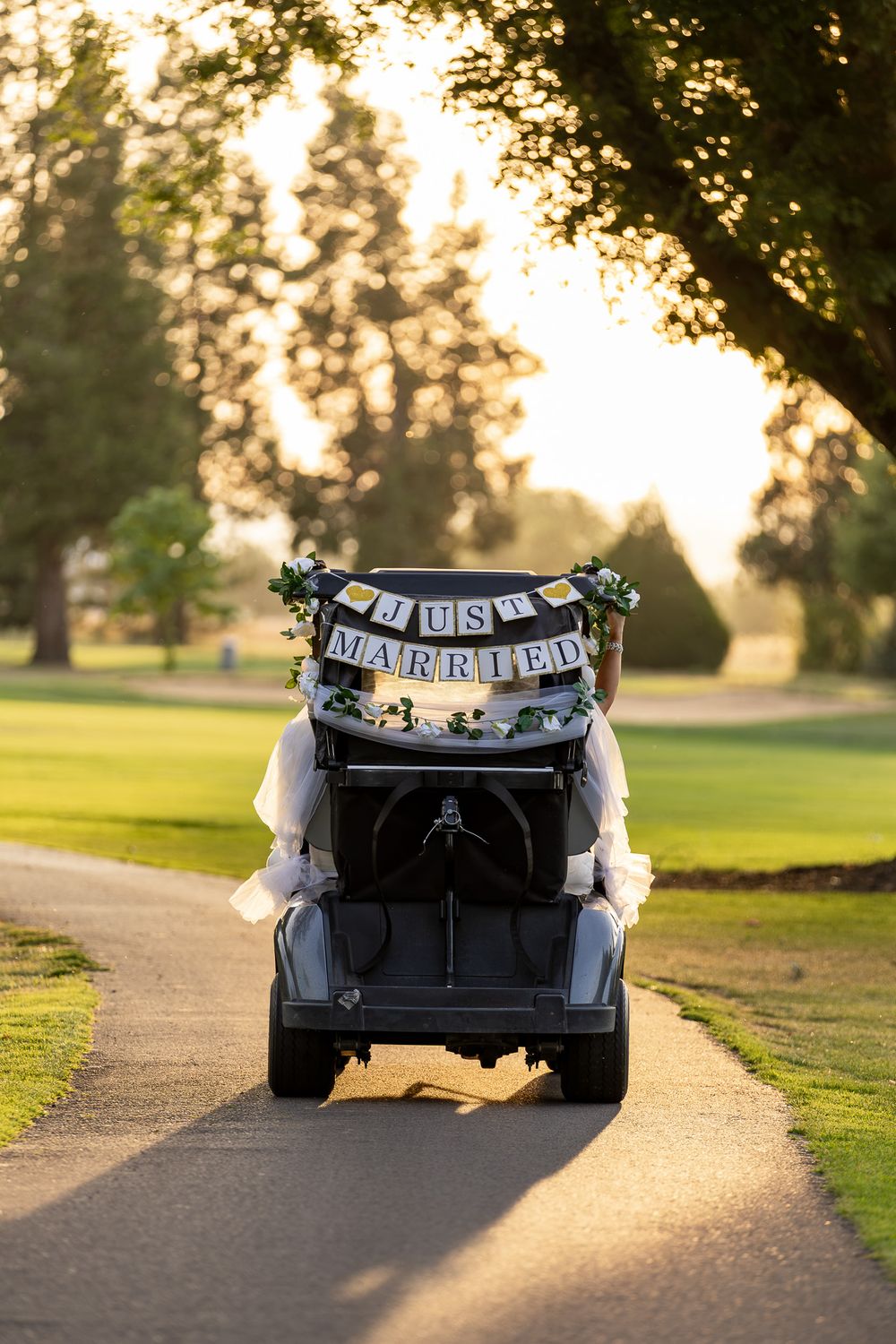 A decorated golf cart with a Just Married banner drives through a sunlit golf course path lined with trees.