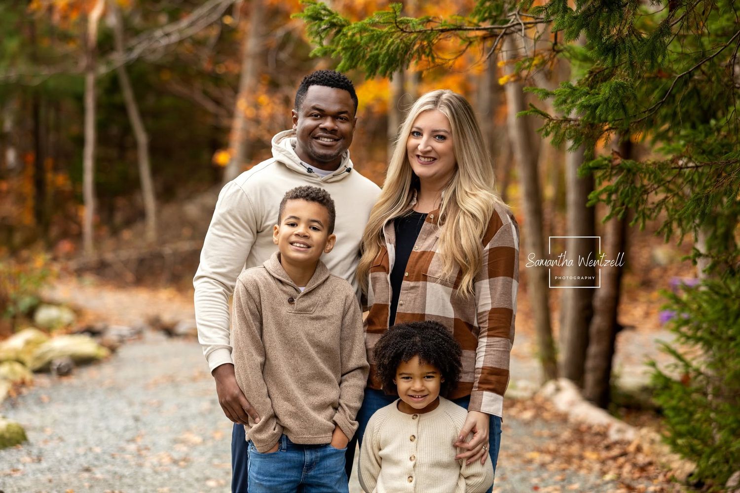 Family of four posing outdoors in autumn forest setting with colorful fall foliage backdrop.