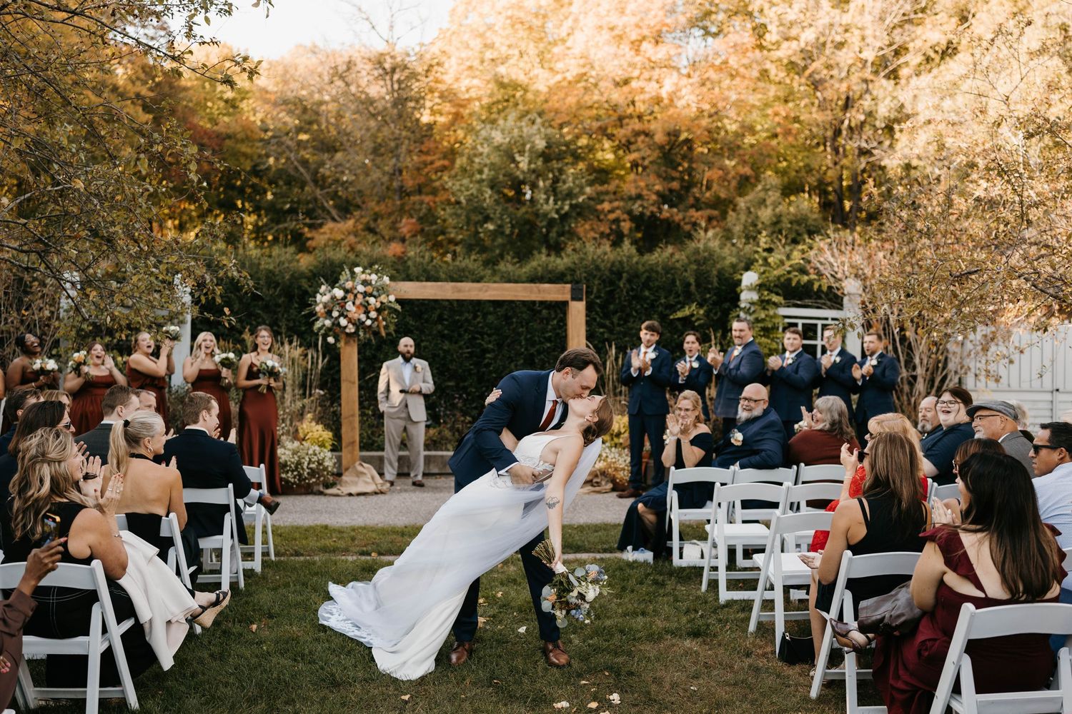 Bride and groom share a romantic dip kiss at outdoor fall wedding ceremony with guests cheering among autumn foliage.