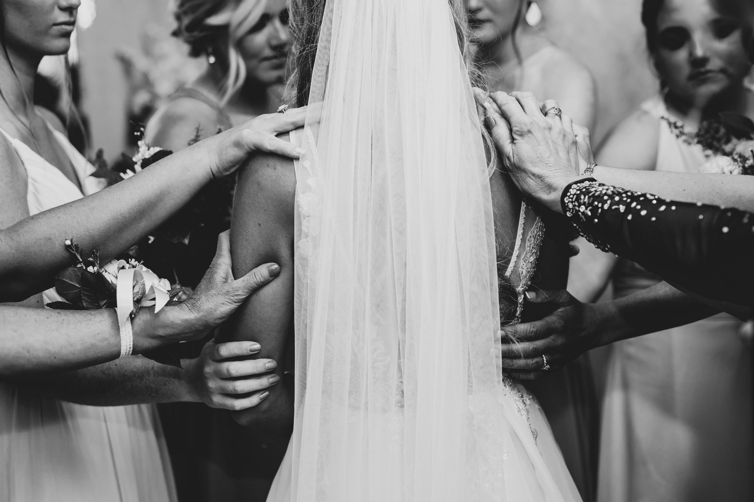 Black and white photo of bride in wedding veil surrounded by bridesmaids placing hands on her shoulders.